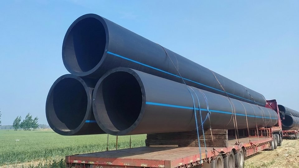 Large-diameter black HDPE pressure pipes with blue stripes loaded for delivery beside a field